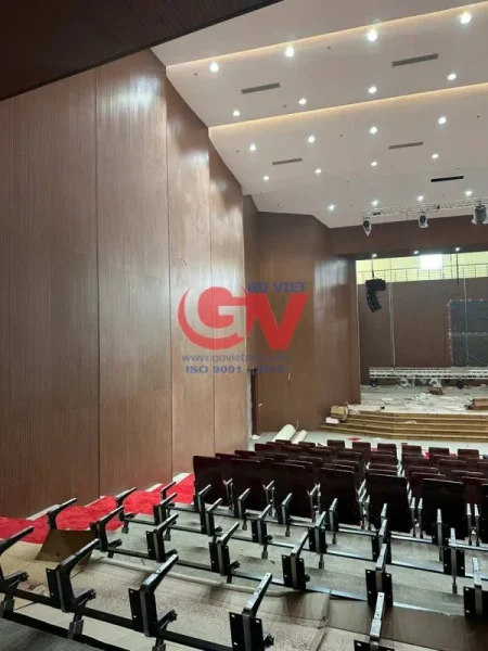Vách ốp vân gỗ màu nâu tại hội trường với hàng ghế khán giả và sân khấu.